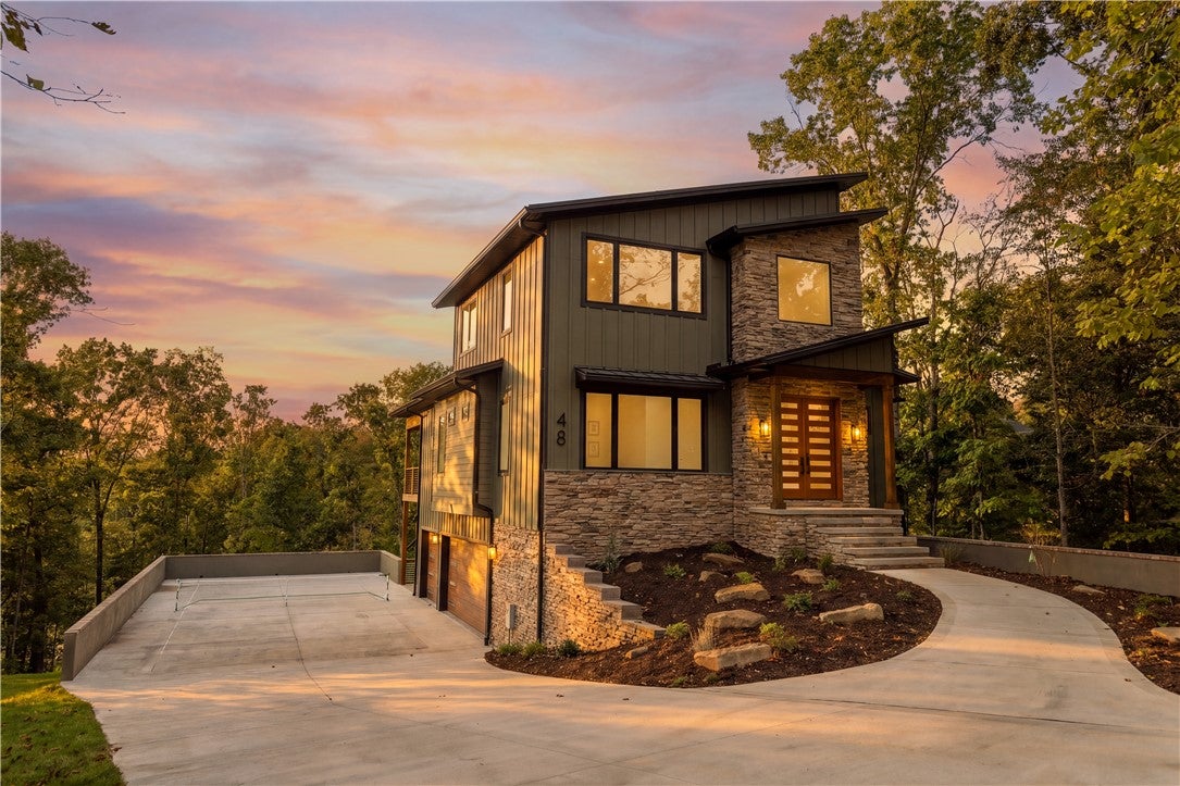 Modern two-story home with stone and olive siding, a concrete driveway, and a tennis court at sunset. [AI-generated text]