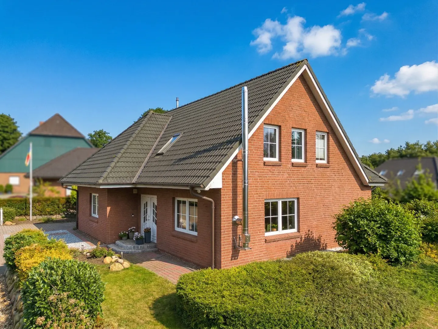 Two-story brick house with a gray roof, white trim, and a silver chimney, surrounded by green bushes and grass under a blue sky.