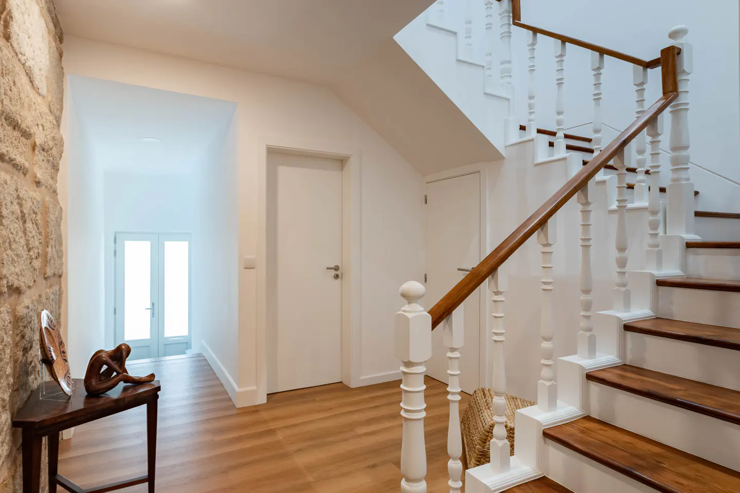 Hallway with wood floors, stone wall, and white staircase with wood railing. A dark wood table with sculpture sits near a doorway.