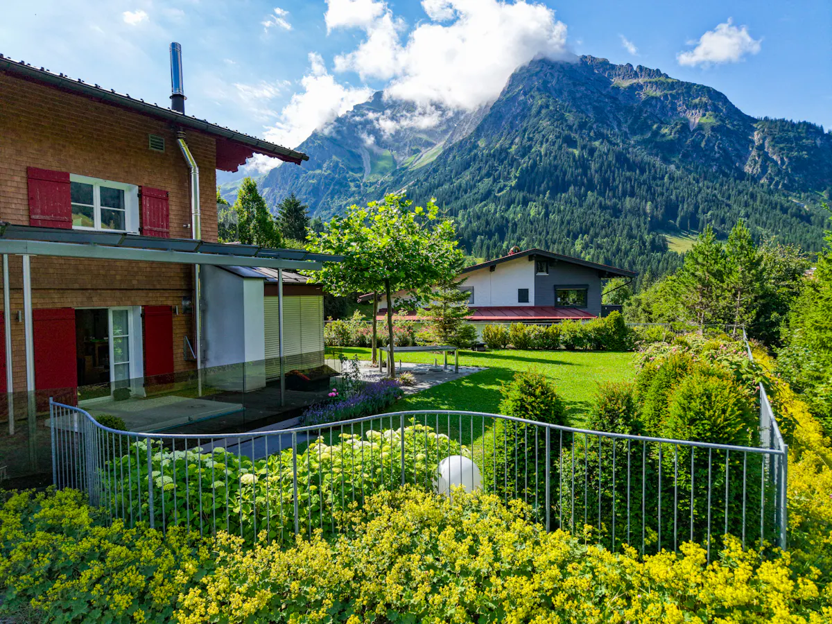 A chalet-style house with red shutters, a green lawn, and a mountain backdrop.
