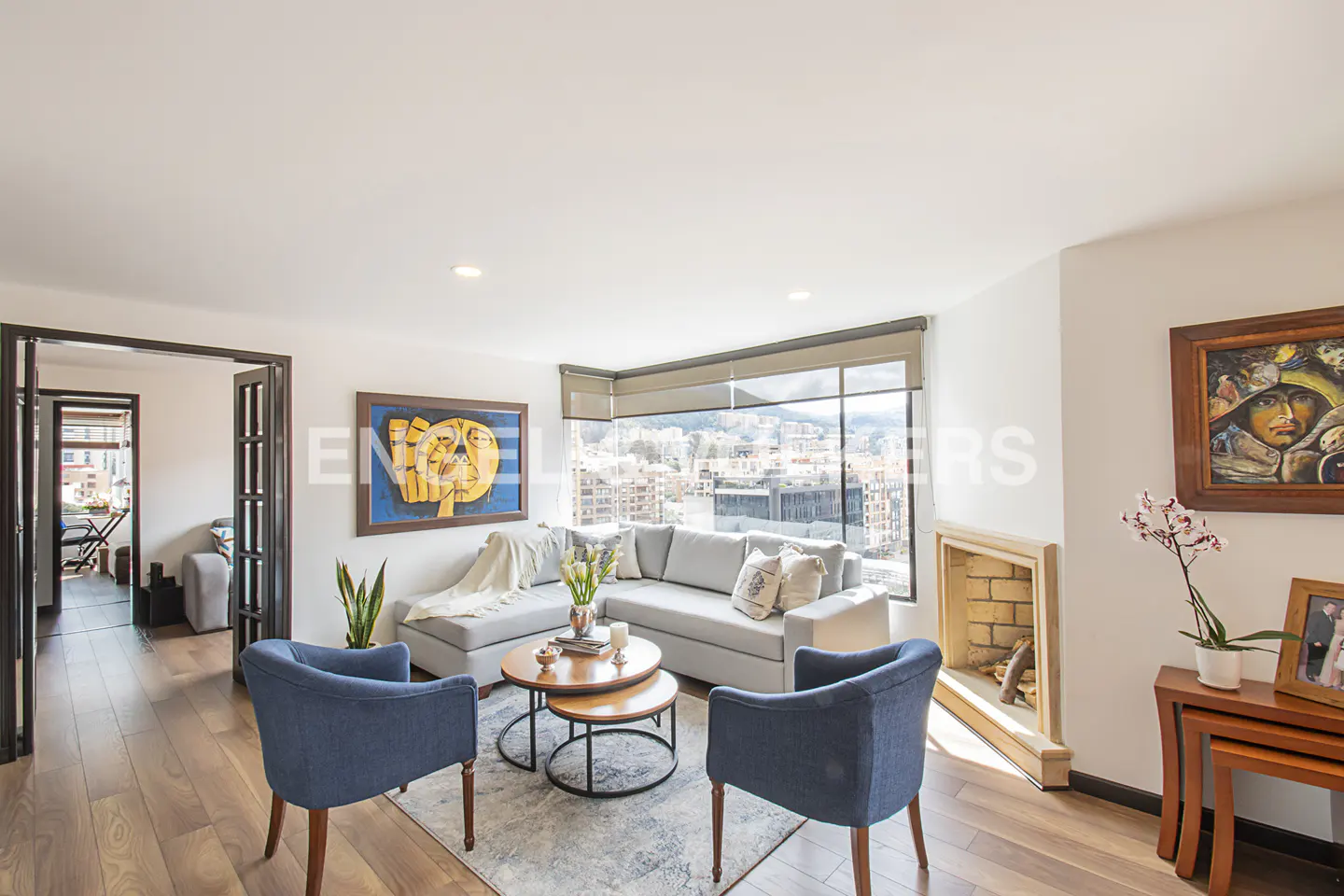 Bright living room with hardwood floors, gray sectional sofa, blue armchairs, and a large window with a city view.