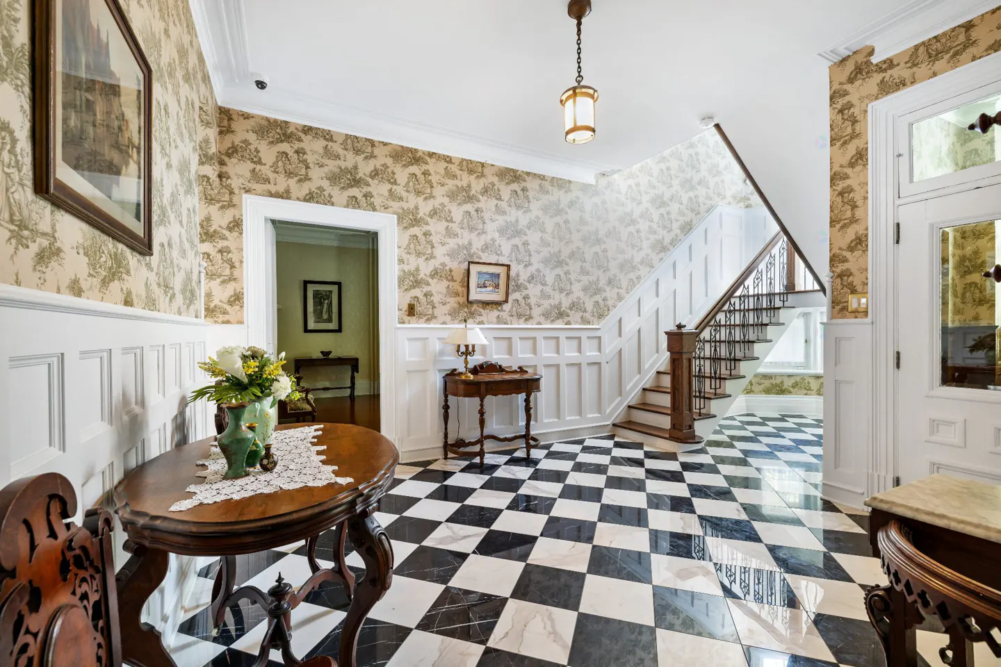 Foyer with black and white checkered floor, staircase, and toile wallpaper. A round table with flowers sits in the foreground.
