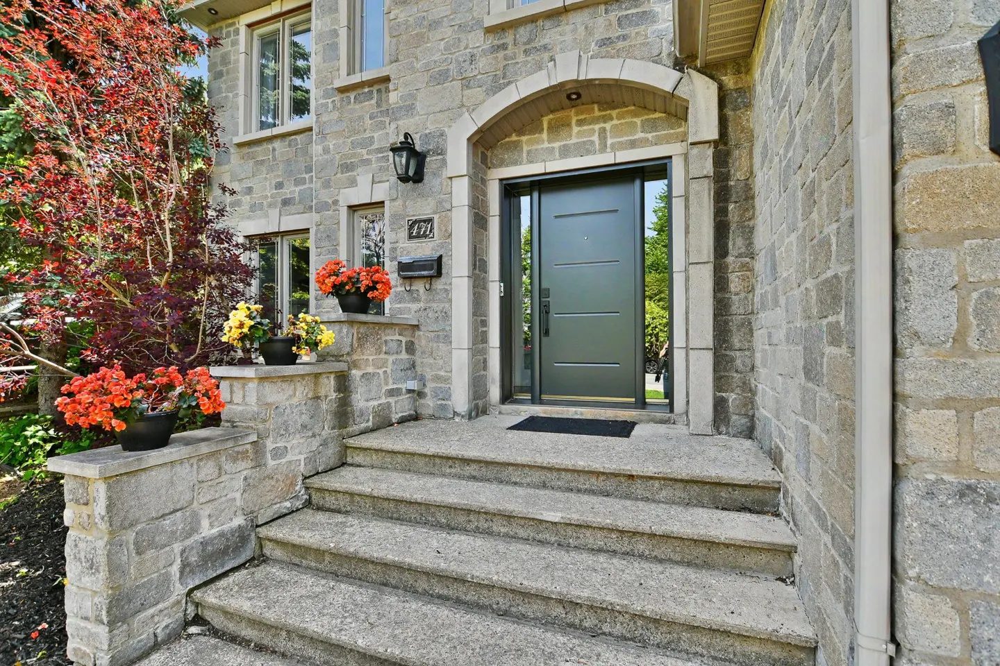 Stone house exterior with concrete steps leading to a modern dark grey front door, accented by vibrant potted flowers.