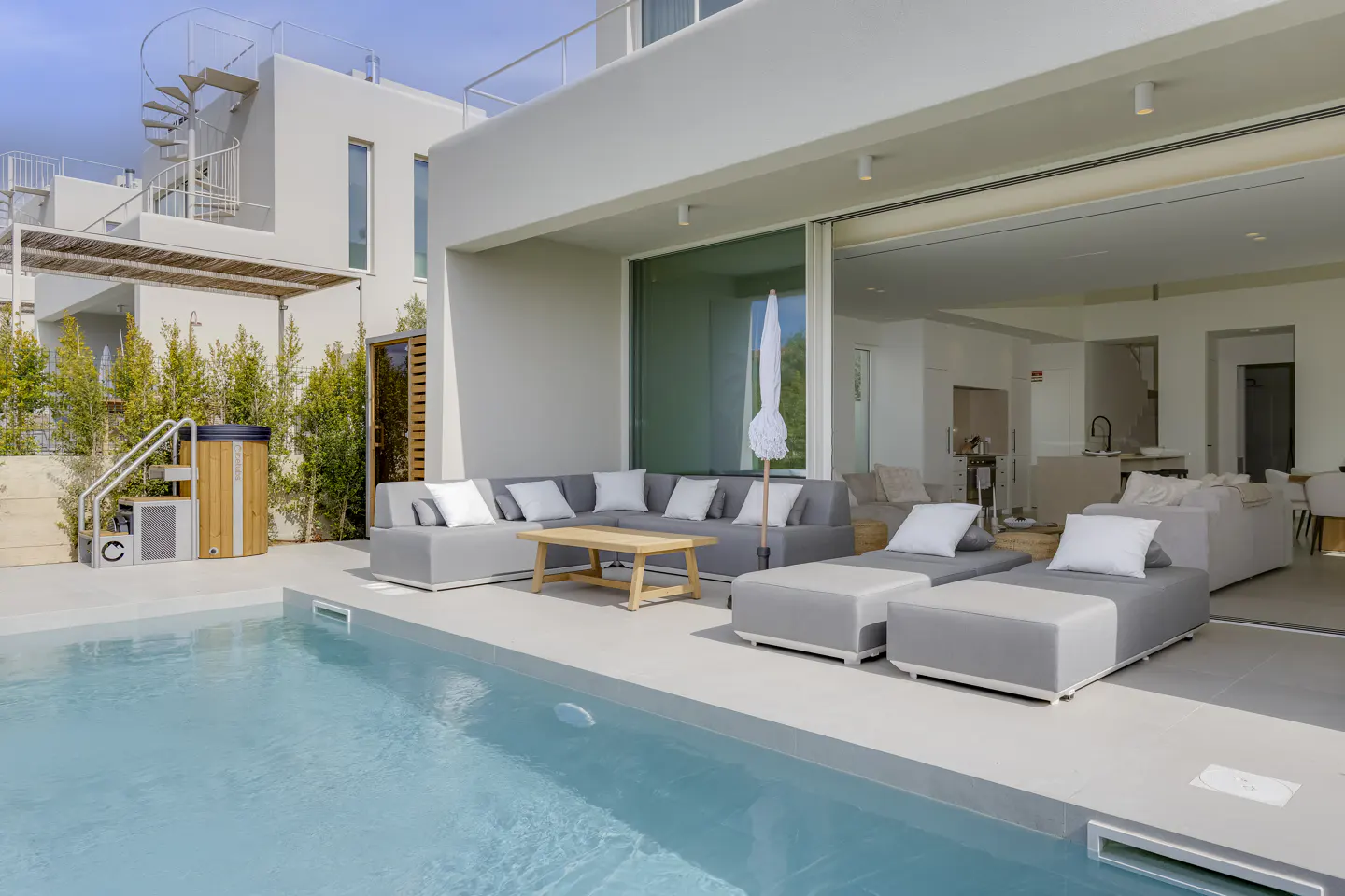 Modern white house with a pool. Gray outdoor furniture sits on the patio. Inside, the living room is visible through sliding glass doors.
