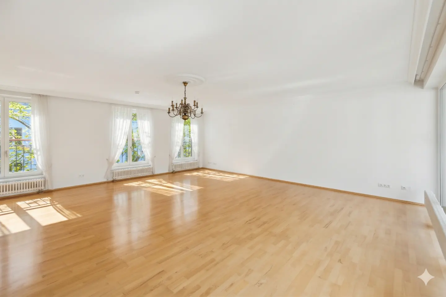 Bright, empty room with hardwood floors, white walls, and a chandelier. Sunlight streams through three windows with sheer white curtains.