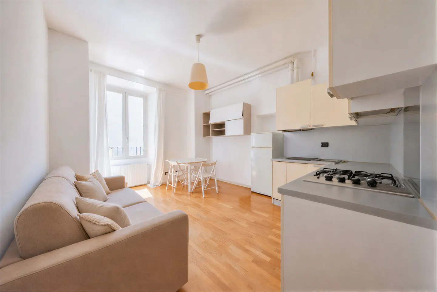 Bright studio apartment with a beige sofa, small dining table, white kitchen cabinets, and light wood flooring.