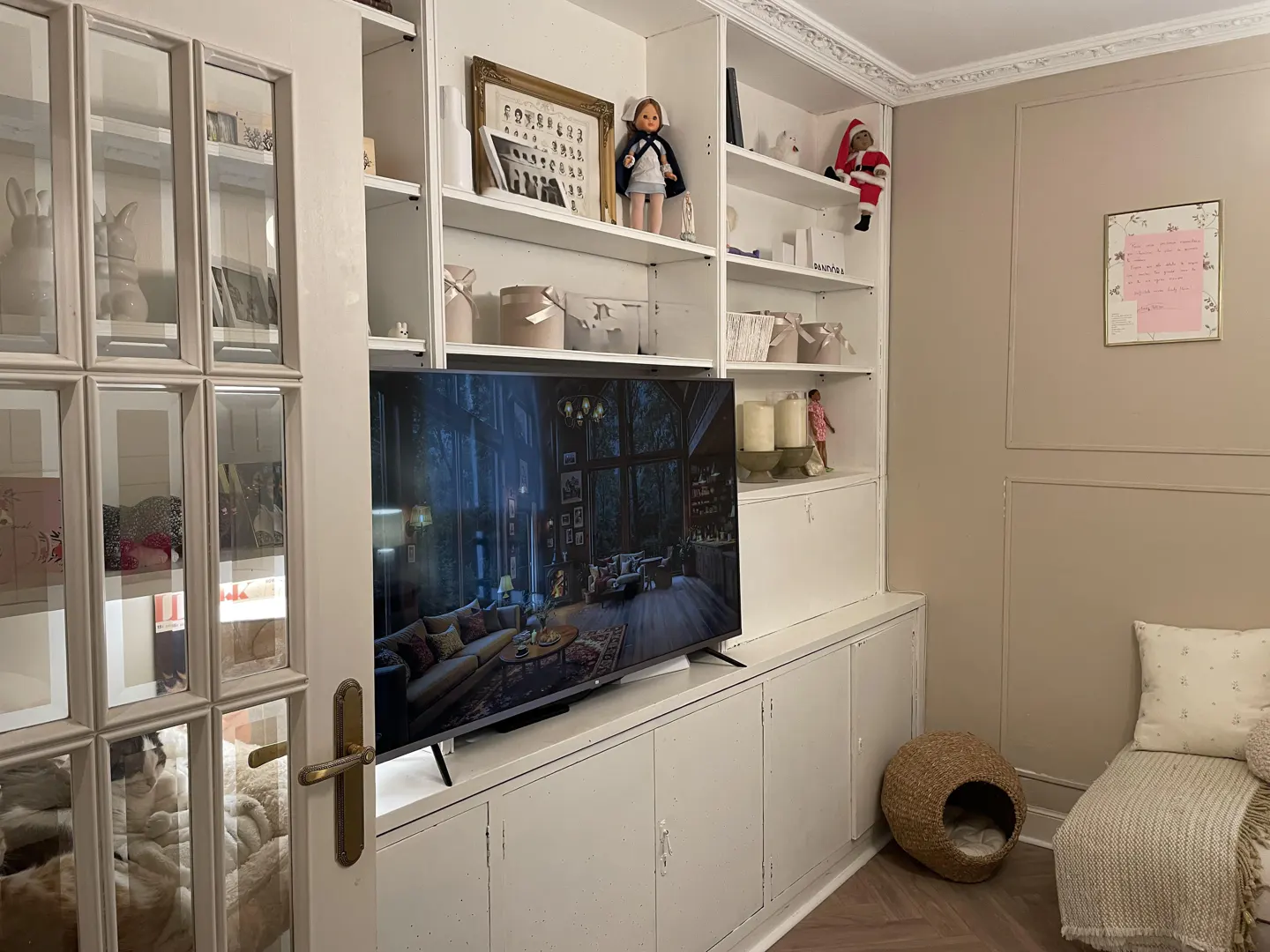 Living room with white built-in shelves, a TV, and a daybed. Shelves are decorated with dolls, photos, and vases. A cat bed sits on the floor.