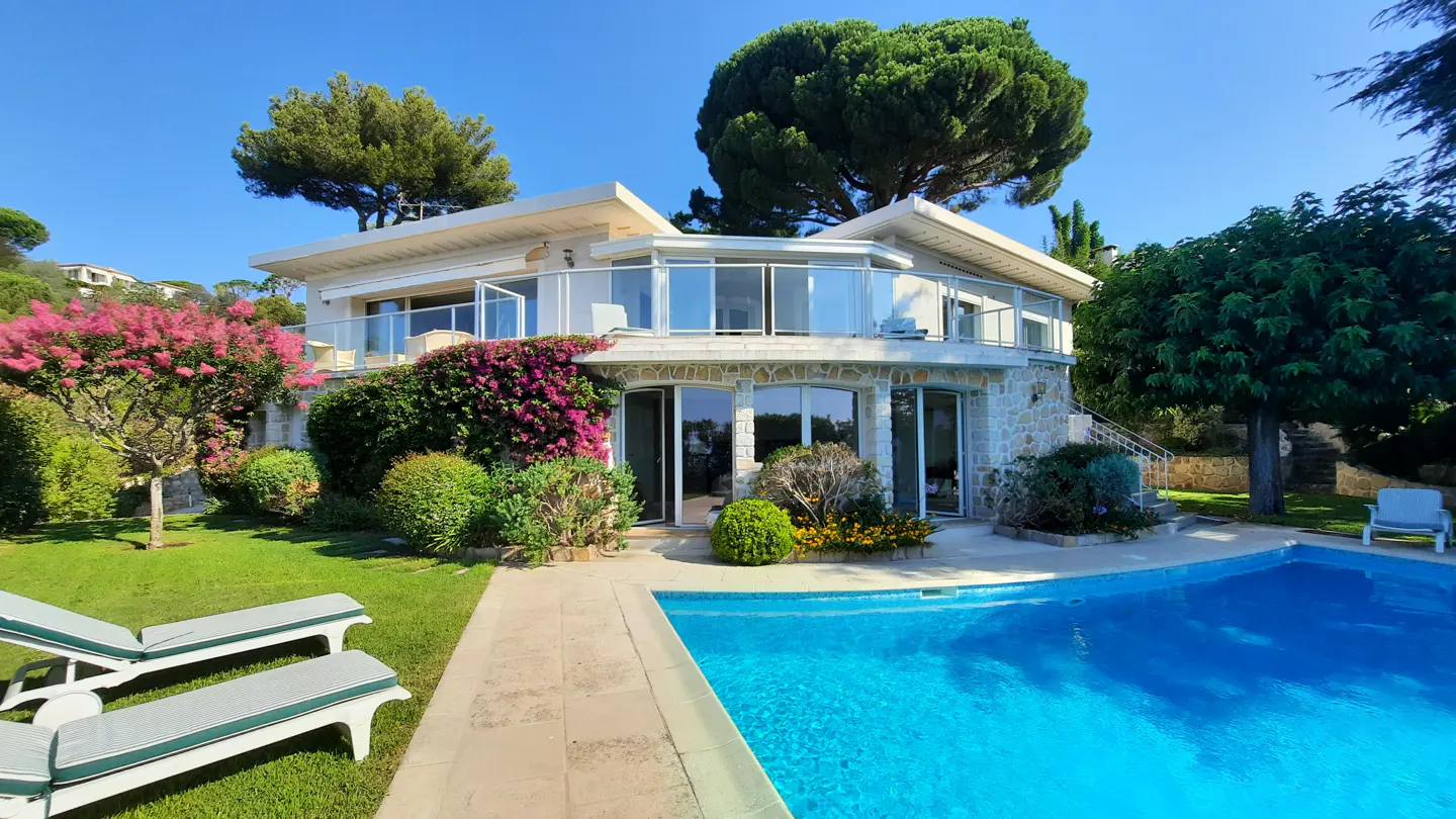 Exterior view of a two-story house with a pool, lawn chairs, and lush landscaping under a clear blue sky.