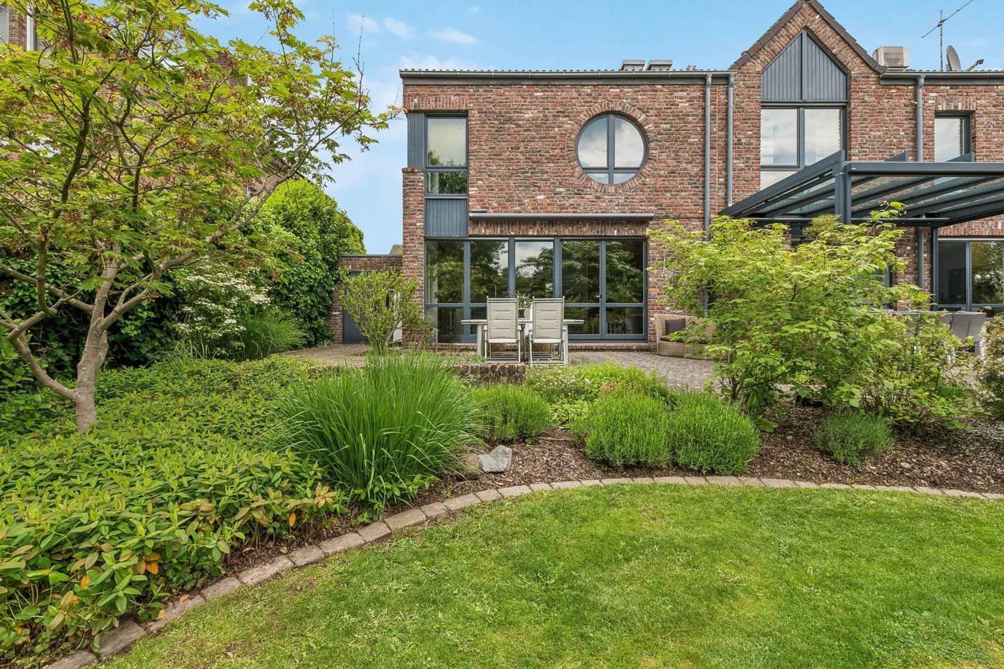 Brick house with a round window, patio with chairs, and a lush green garden.
