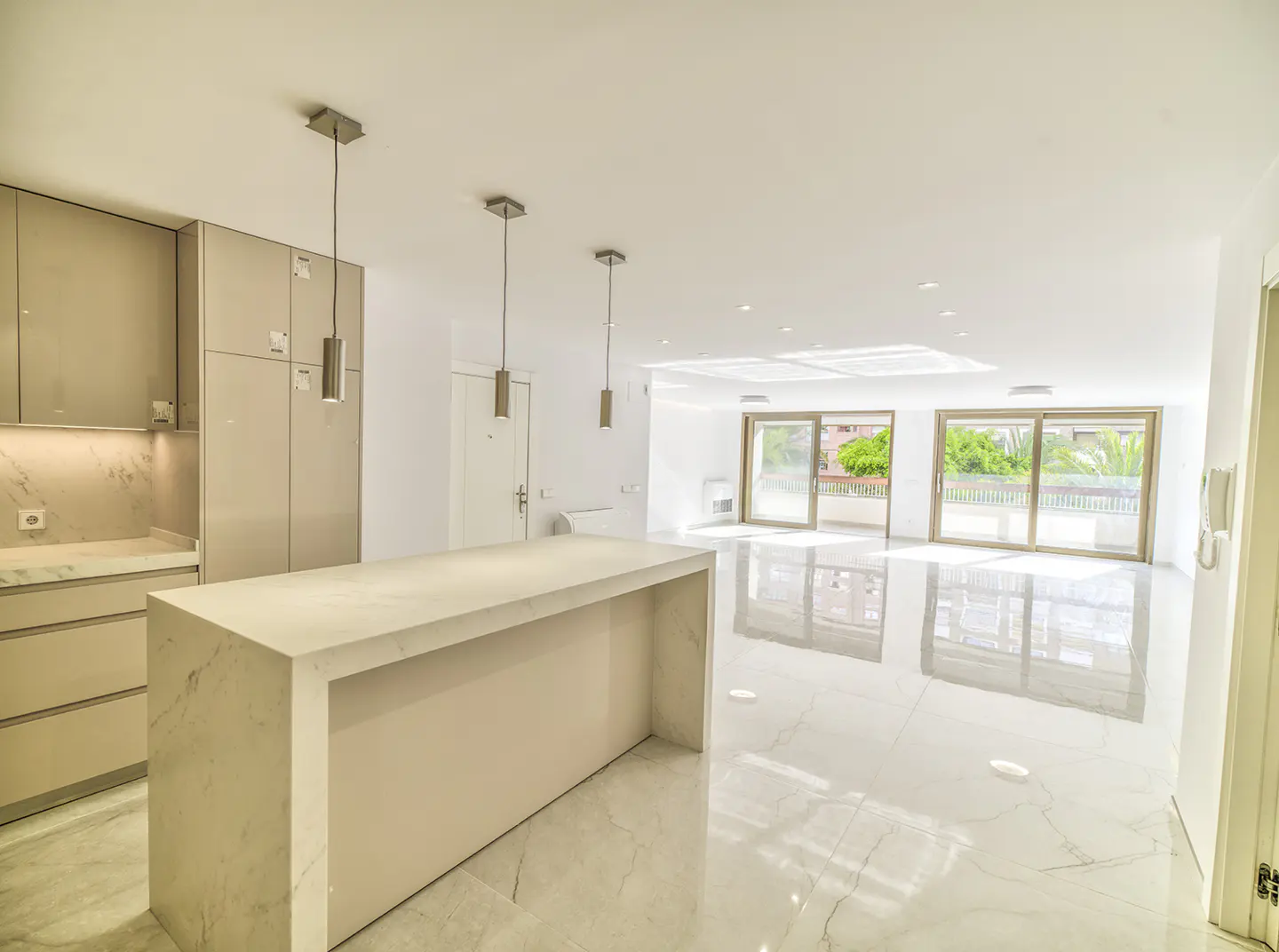 Bright, modern kitchen with marble island, cabinets, and floors. Sliding glass doors lead to a balcony with greenery visible outside.