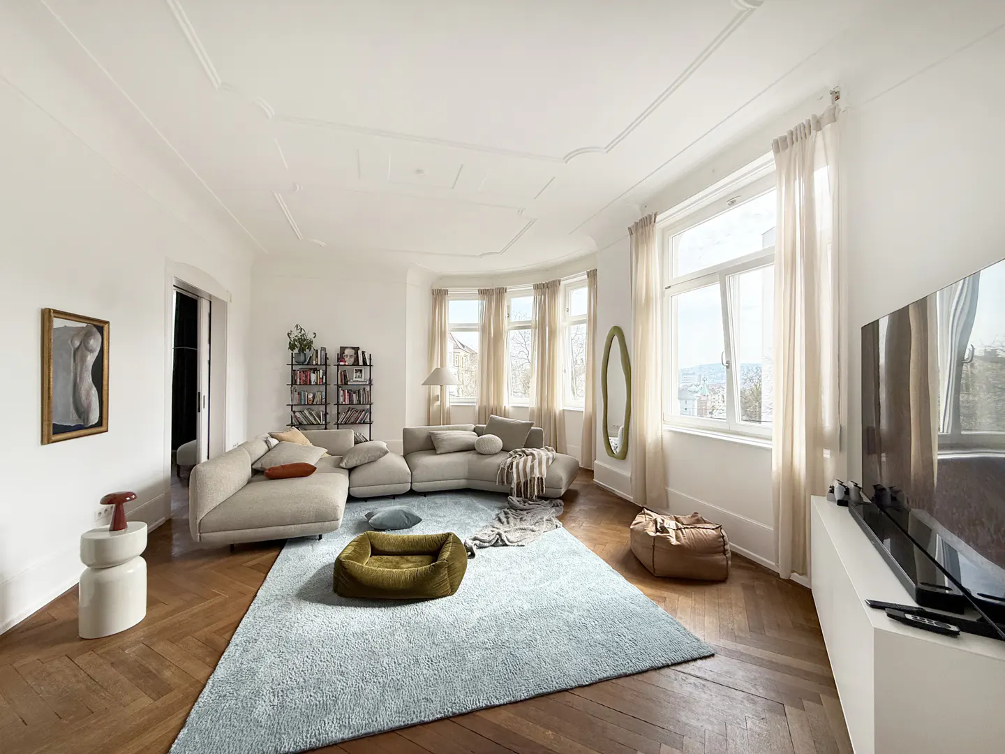 Bright living room with white walls, herringbone wood floors, and a large light blue rug. A beige sectional sofa faces a TV. Large windows with sheer curtains let in natural light.