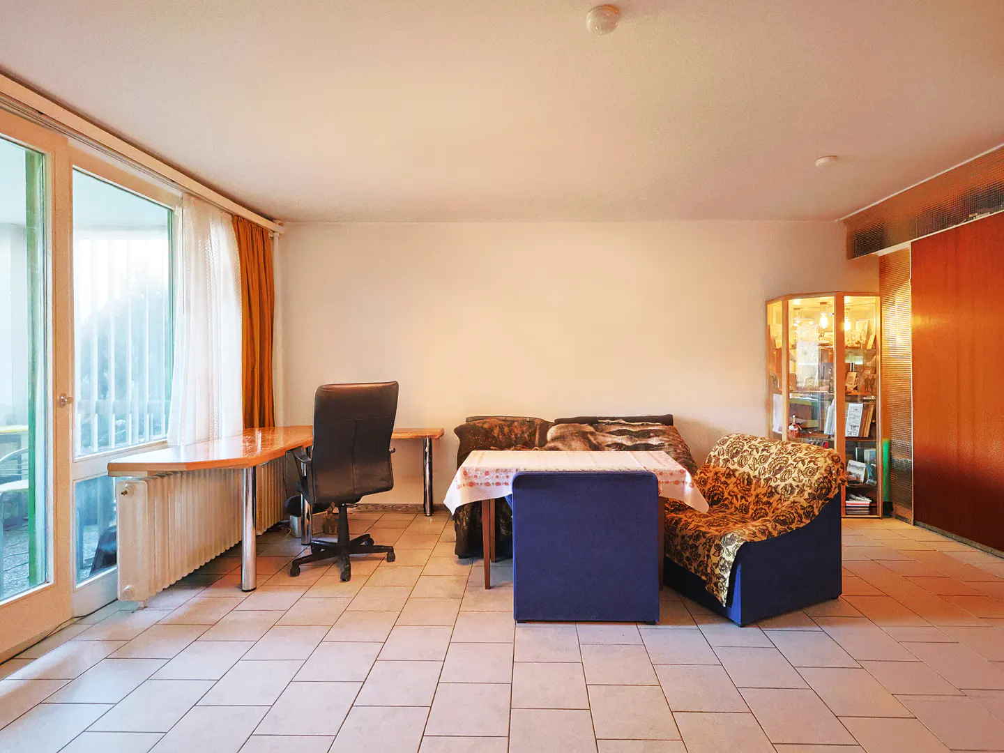 Bright room with tile floor, desk, chair, sofa, and blue chair. A glass cabinet stands against the wall.