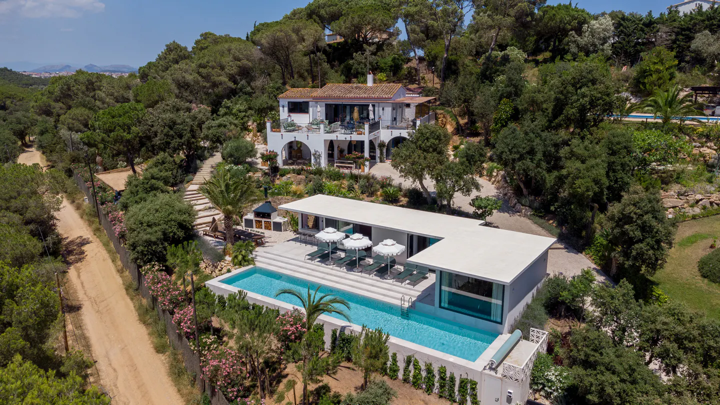 Aerial view of a white two-story house with a pool, umbrellas, and lounge chairs surrounded by trees.