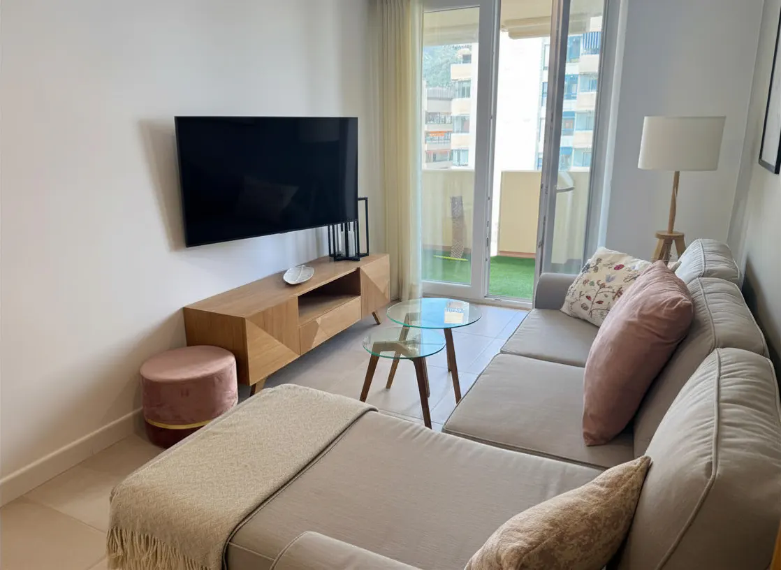 Living room with a gray sectional sofa, TV, wood cabinet, glass tables, and a balcony with a view of buildings.