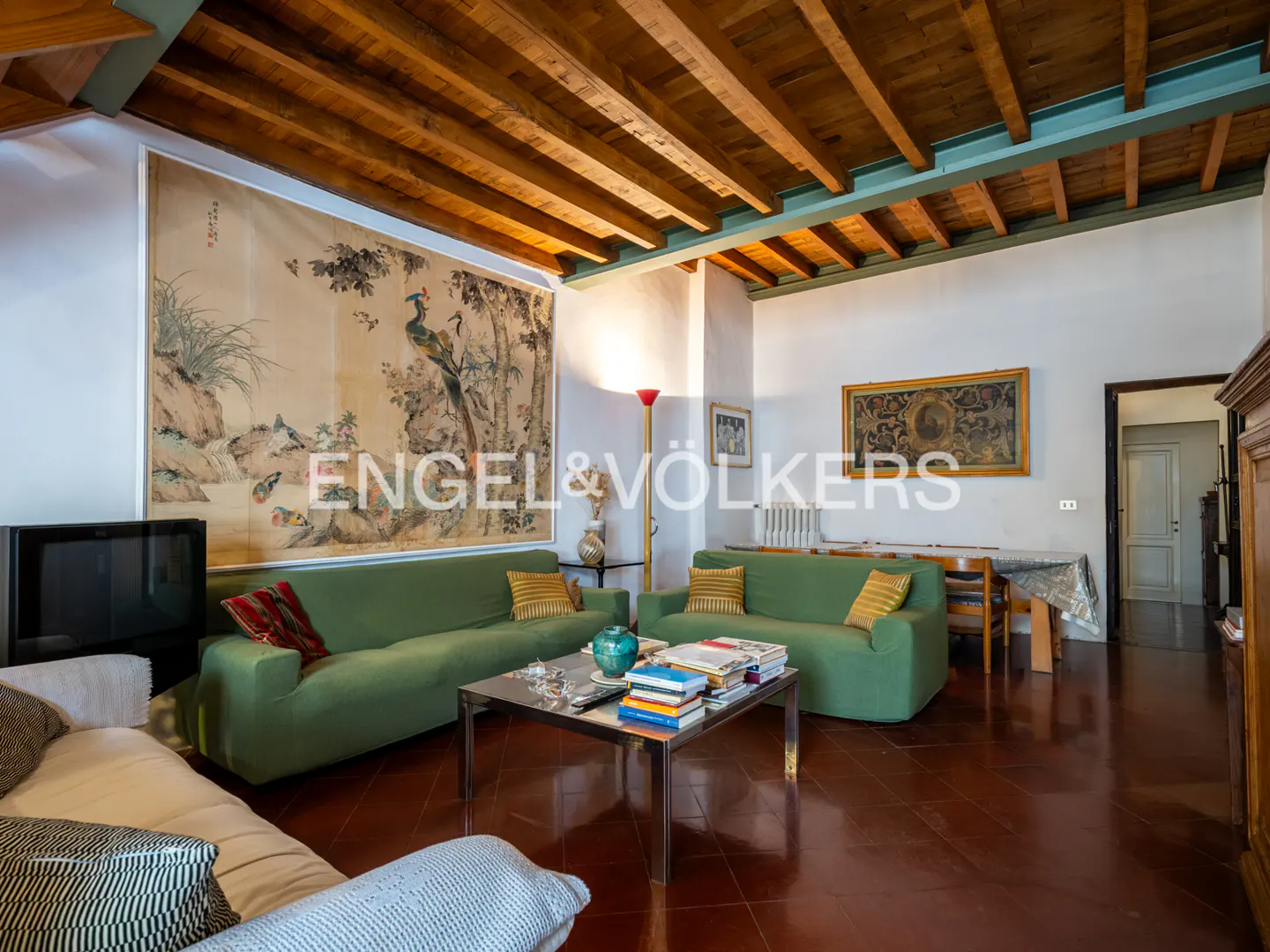 Living room with green sofas, a coffee table with books, and a large Asian-style tapestry on the wall. Wooden beamed ceiling.