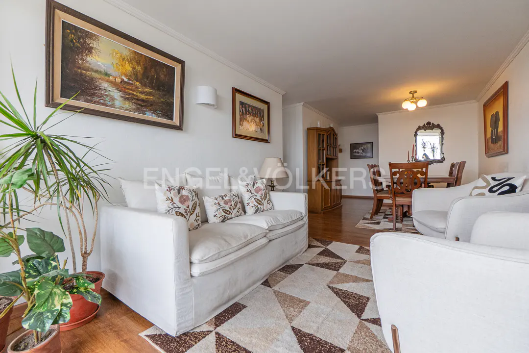 Bright living room with white sofas, patterned rug, potted plant, and a dining area with wooden furniture in the background.