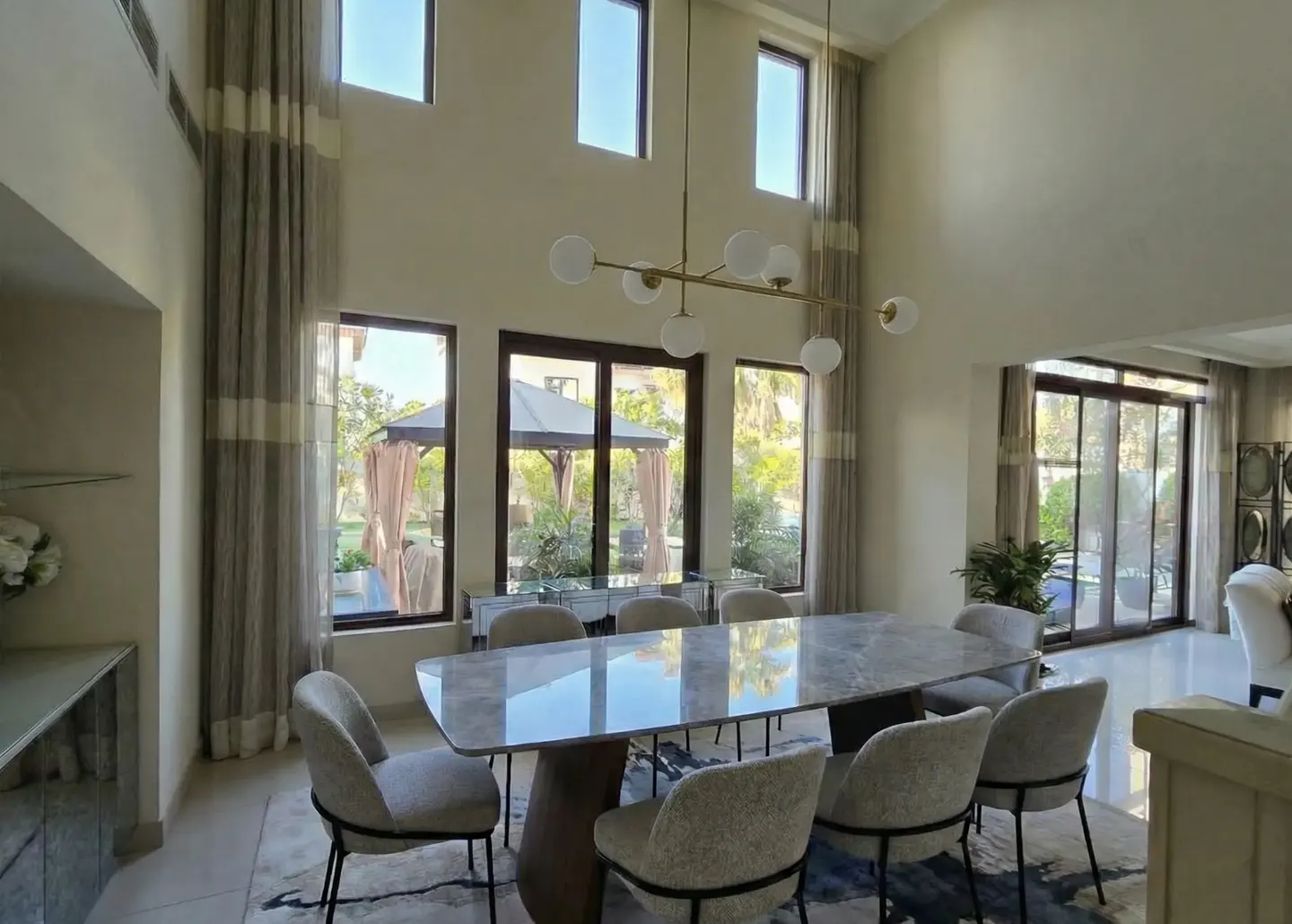 Bright dining room with a marble table, upholstered chairs, and a modern chandelier. Large windows offer a view of the outdoor patio.
