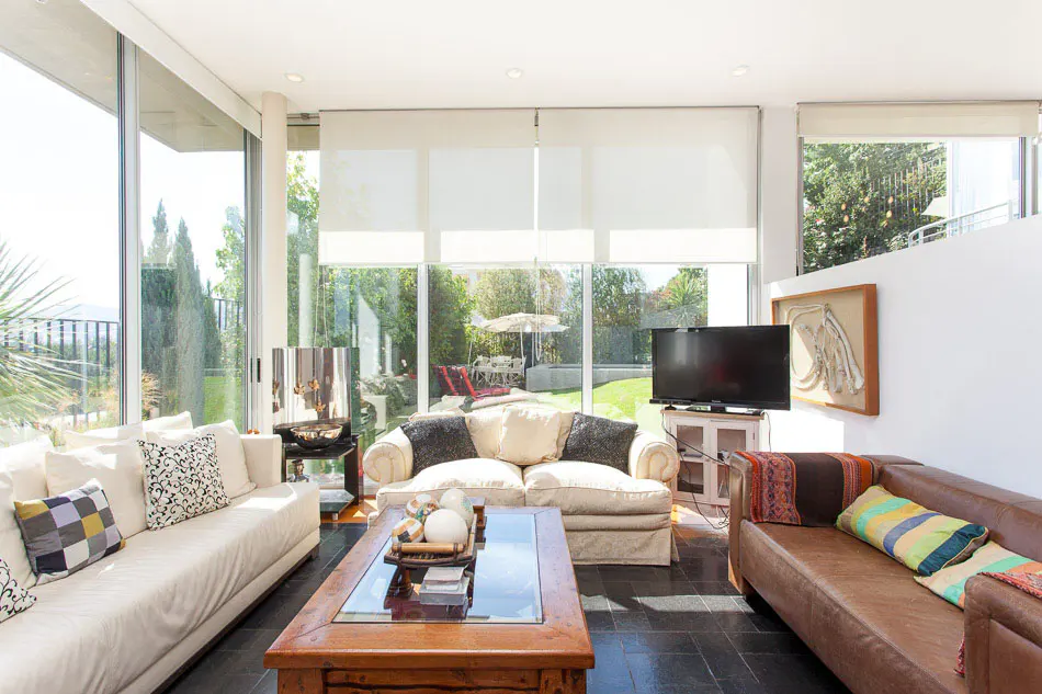 Bright living room with white and brown sofas, a wooden coffee table, and large windows overlooking a green yard.