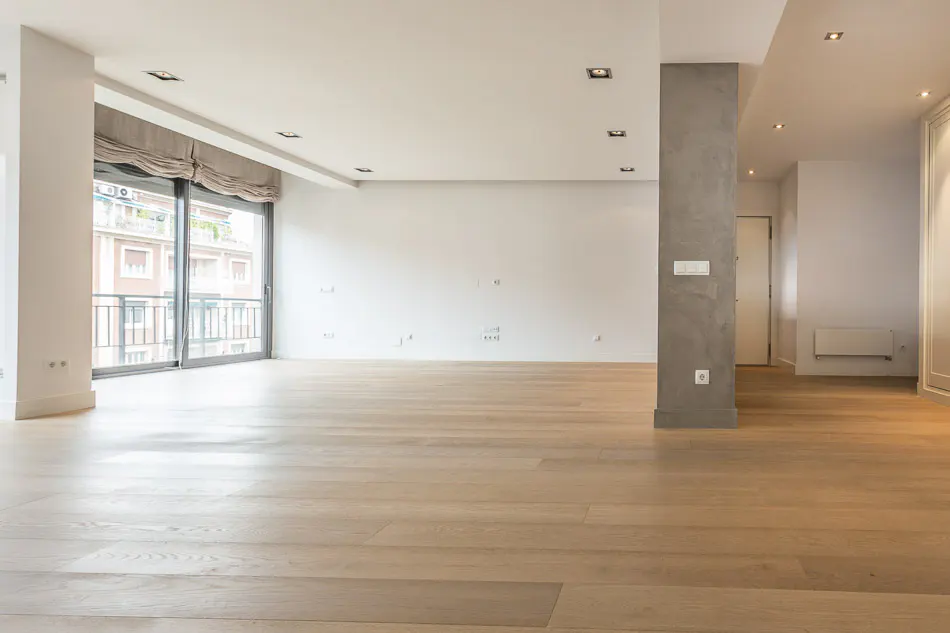 Bright, empty room with light wood floors, white walls, and recessed lighting. Large window with gray curtains shows a city view. Gray pillar near a doorway.