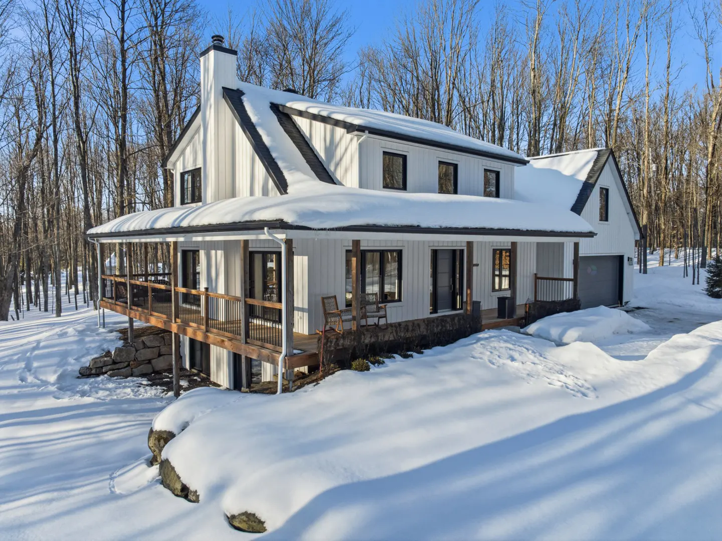 A white farmhouse with black trim and a snow-covered roof sits in a snowy landscape. A wooden porch wraps around the house.