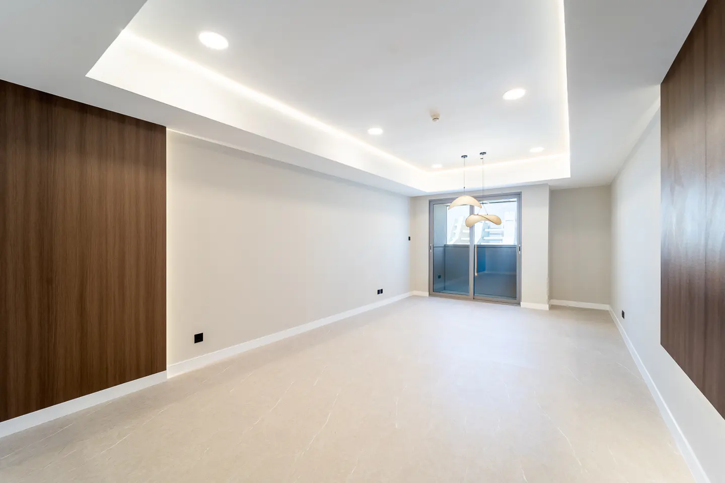 Bright, empty room with wood paneling, white walls, and tile floor. Sliding glass doors lead to a balcony. Modern pendant lights hang from the ceiling.