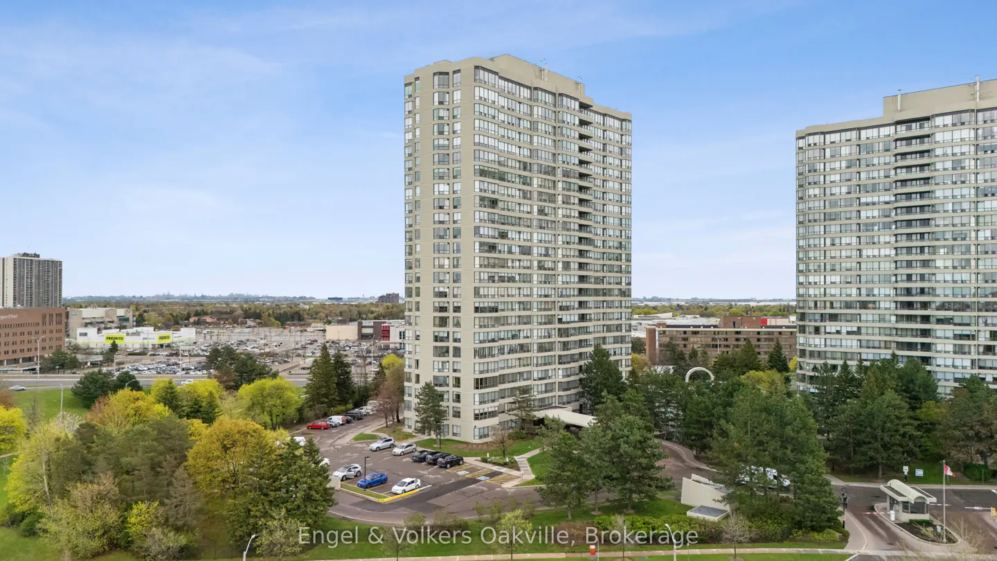 Exterior shot of a tall, light gray condo building with many windows, surrounded by green trees and a parking lot.