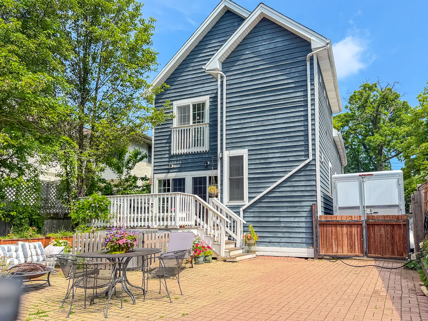 Blue two-story house with a white deck, patio furniture, and a brick-paved backyard on a sunny day.