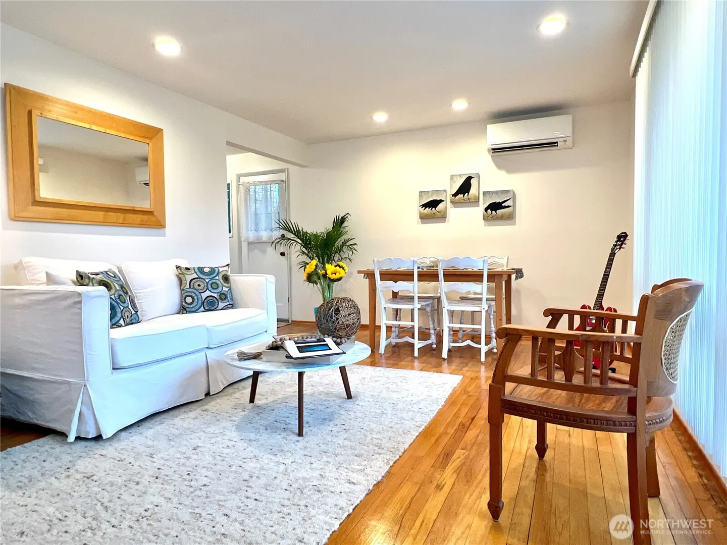 Bright living room with white sofa, round marble table, dining set, and wooden chair on hardwood floors.