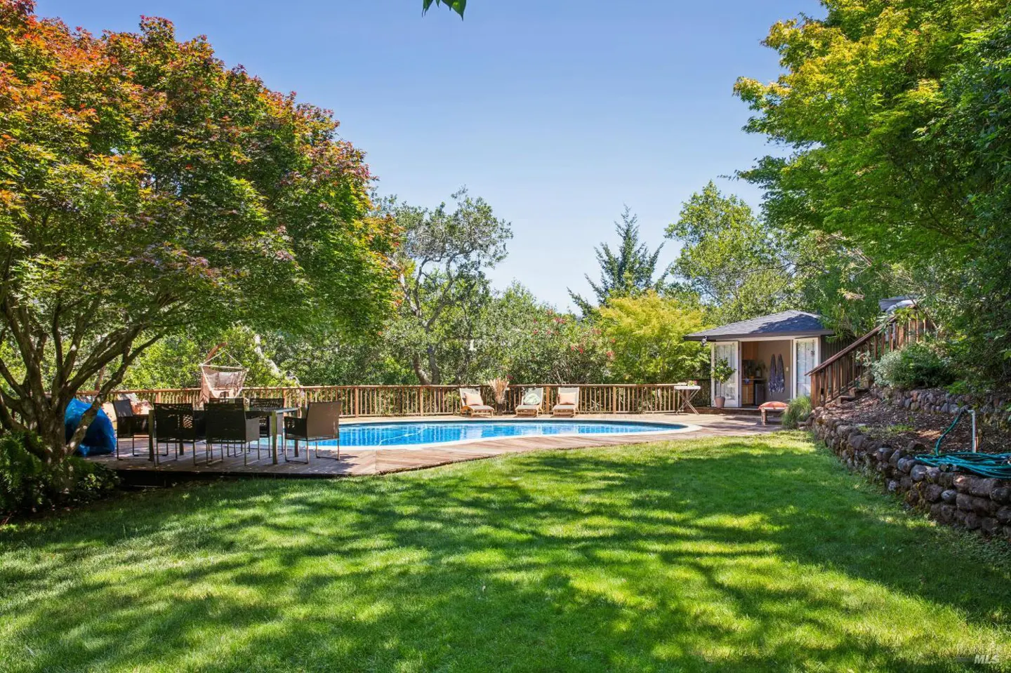 Backyard with a blue pool, wooden deck, table, chairs, and a small pool house surrounded by green trees and grass.