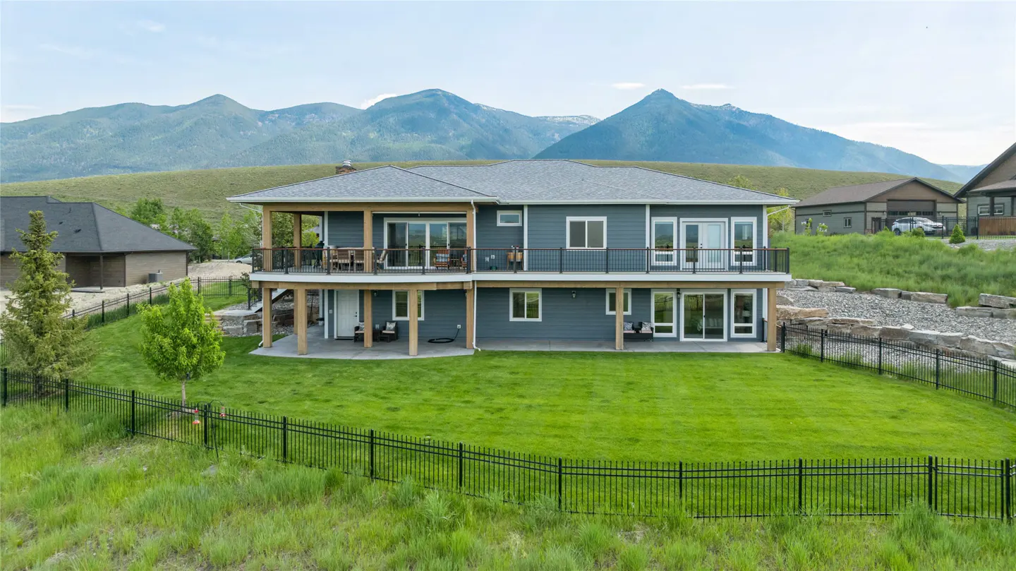 Two-story blue house with a gray roof, a large green lawn, and a black metal fence. Mountains are visible in the background.