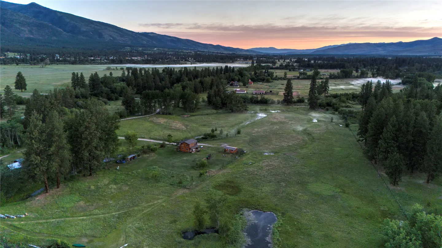 Aerial view of a log cabin home in a green meadow with mountains and a pink sky in the background.