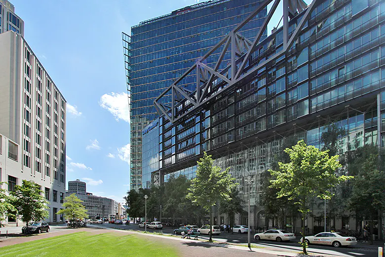 Modern city view with glass buildings, green trees, and parked cars under a blue sky.