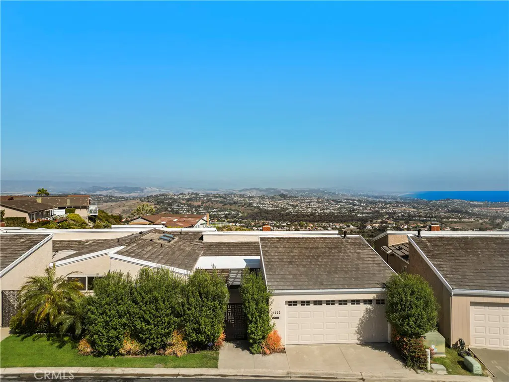Aerial view of a beige single-story house with a gray roof, green bushes, and a view of the city and ocean.