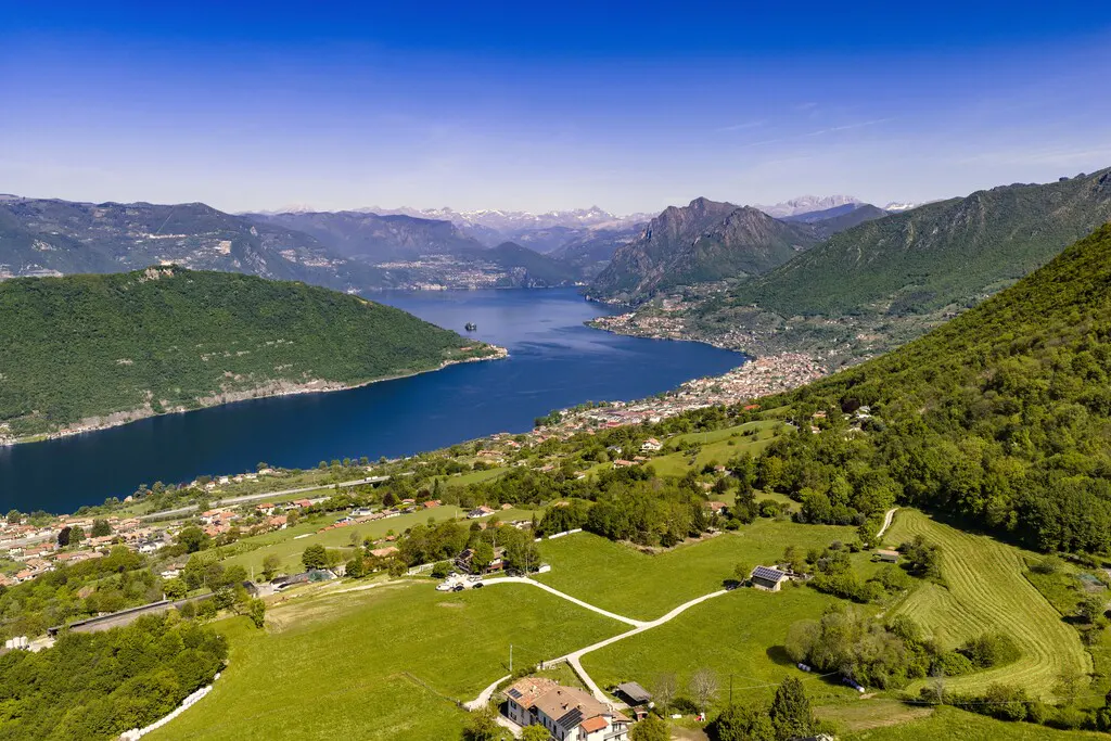Scenic aerial view of Lake Iseo, Italy, with green hills, a blue lake, and a clear blue sky. A small town lines the shore.
