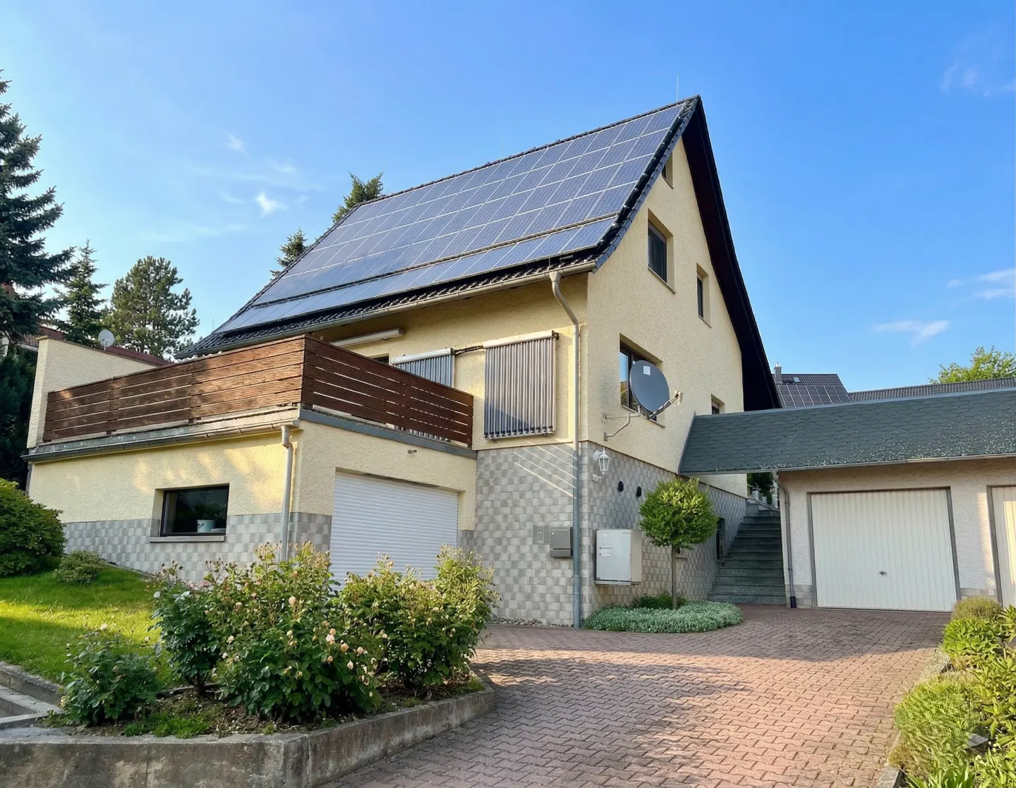 Two-story beige house with solar panels on the roof, a balcony, and a brick driveway.