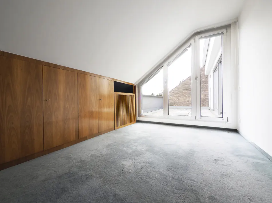 Attic room with blue carpet, wood cabinets, and a large triangular window with a view of a brick building.