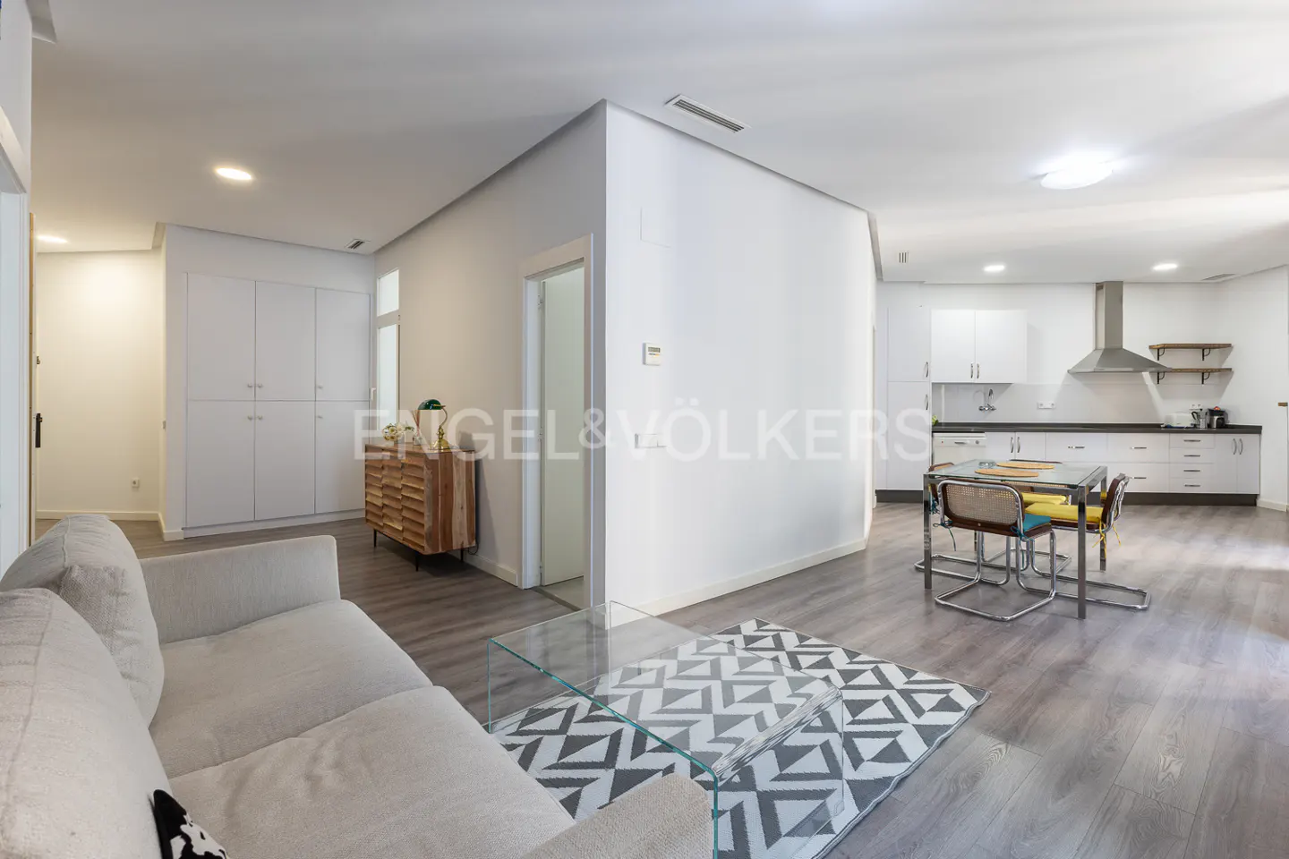 Bright, open-concept apartment with white walls, gray wood floors, and a modern kitchen. A cream sofa and glass table sit in the foreground.