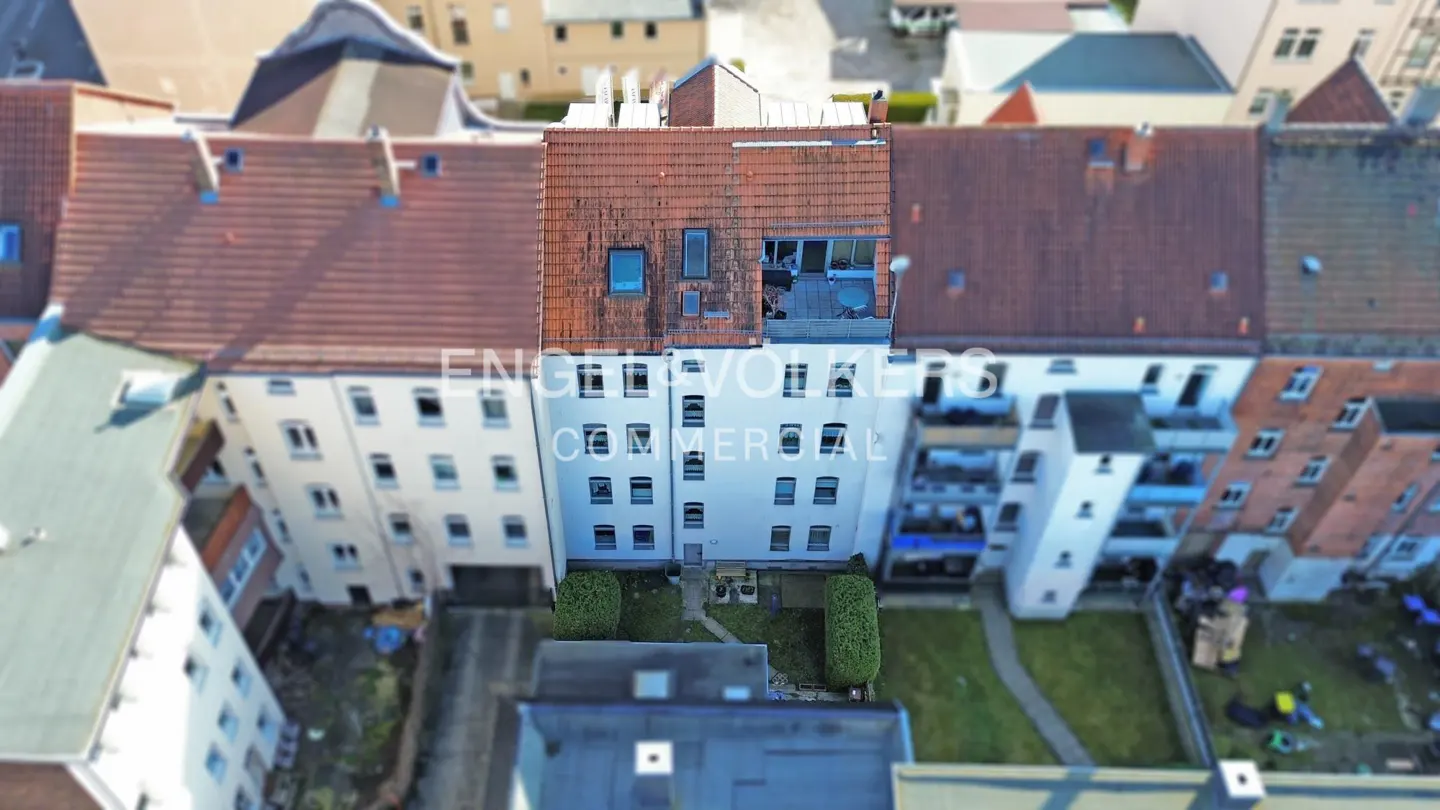 Aerial view of a white apartment building with a red tile roof, surrounded by other buildings and greenery.