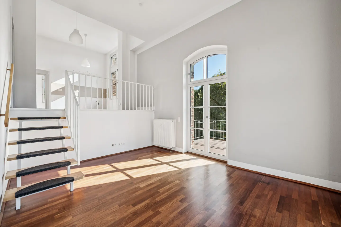 Bright, modern living room with hardwood floors, white walls, and a staircase leading to a loft area. Sunlight streams through a large arched window.