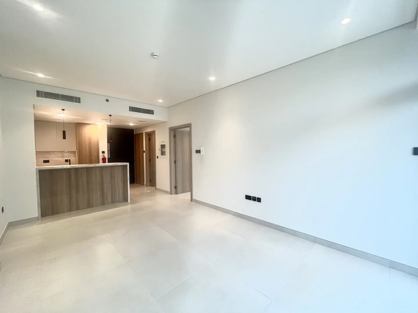 Bright, empty apartment with white walls and tile floors. A kitchen with wood cabinets is visible in the background.