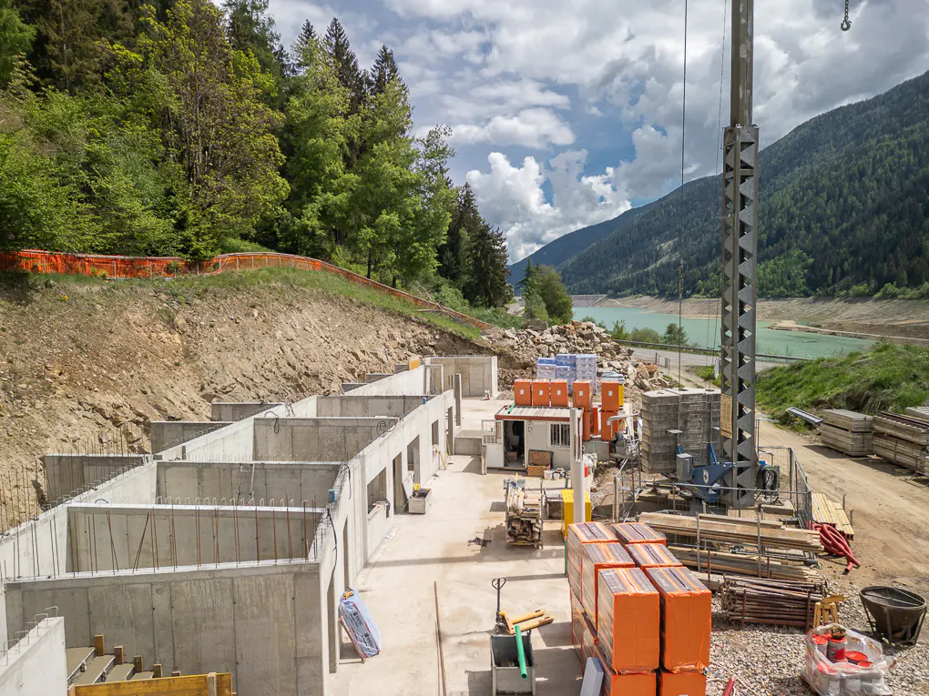 Construction site with concrete foundations, a crane, and building materials. Mountains and a lake are visible in the background.
