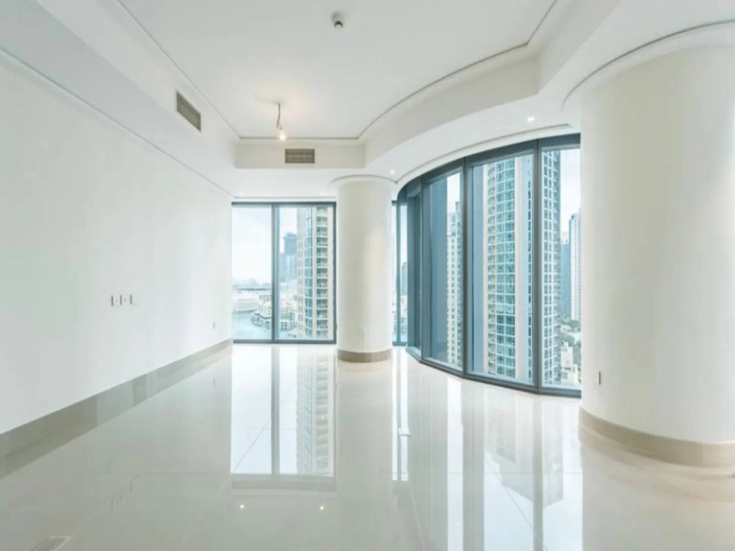 Bright, empty room with white walls, glossy tile floor, and floor-to-ceiling windows. Cityscape visible through the windows.