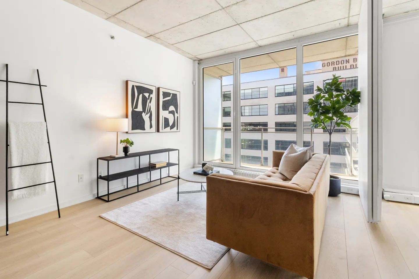 Living room with a tan sofa, black metal shelving, and ladder with a white towel. Large windows overlook a building.