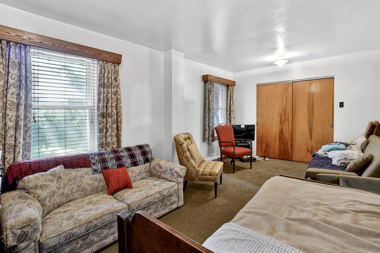 A living room with a floral sofa, a tan armchair, a red accent chair, brown carpet, and wooden sliding closet doors.