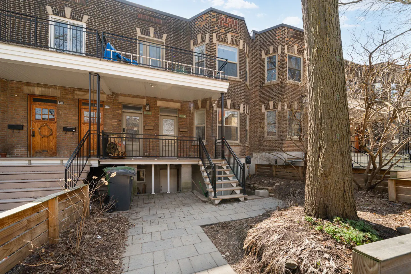 Brick row houses with black iron railings and wooden doors. A stone patio leads to stairs. A large tree is on the right.