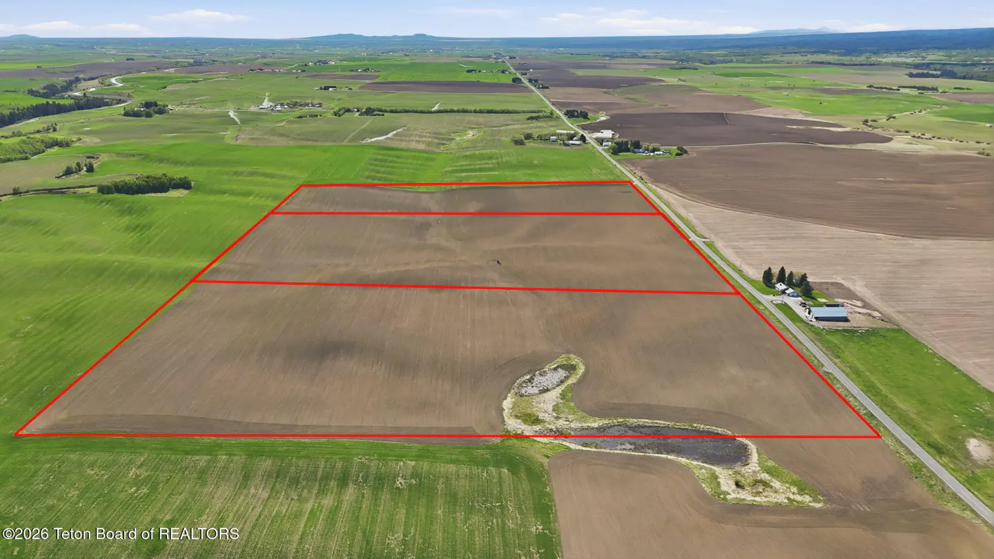 Aerial view of a large, brown, rectangular plot of land outlined in red, surrounded by green fields and a road with a house.