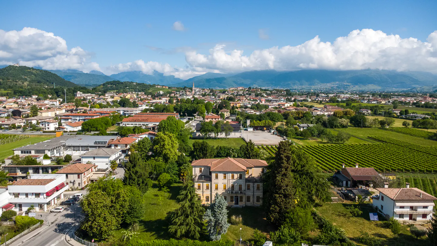 Aerial view of a town with red-roofed buildings, green trees, and vineyards, set against a backdrop of mountains and a blue sky with white clouds.