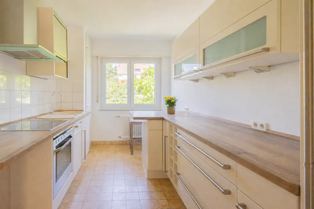 Bright kitchen with white cabinets, wood countertops, and tile floor. A window overlooks greenery. A small table sits near the window.