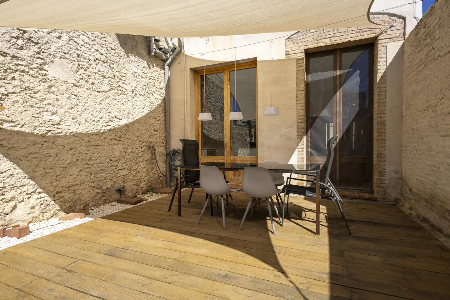 Outdoor patio with wooden deck, table, and chairs under a shade sail. Stone walls and doors in the background.