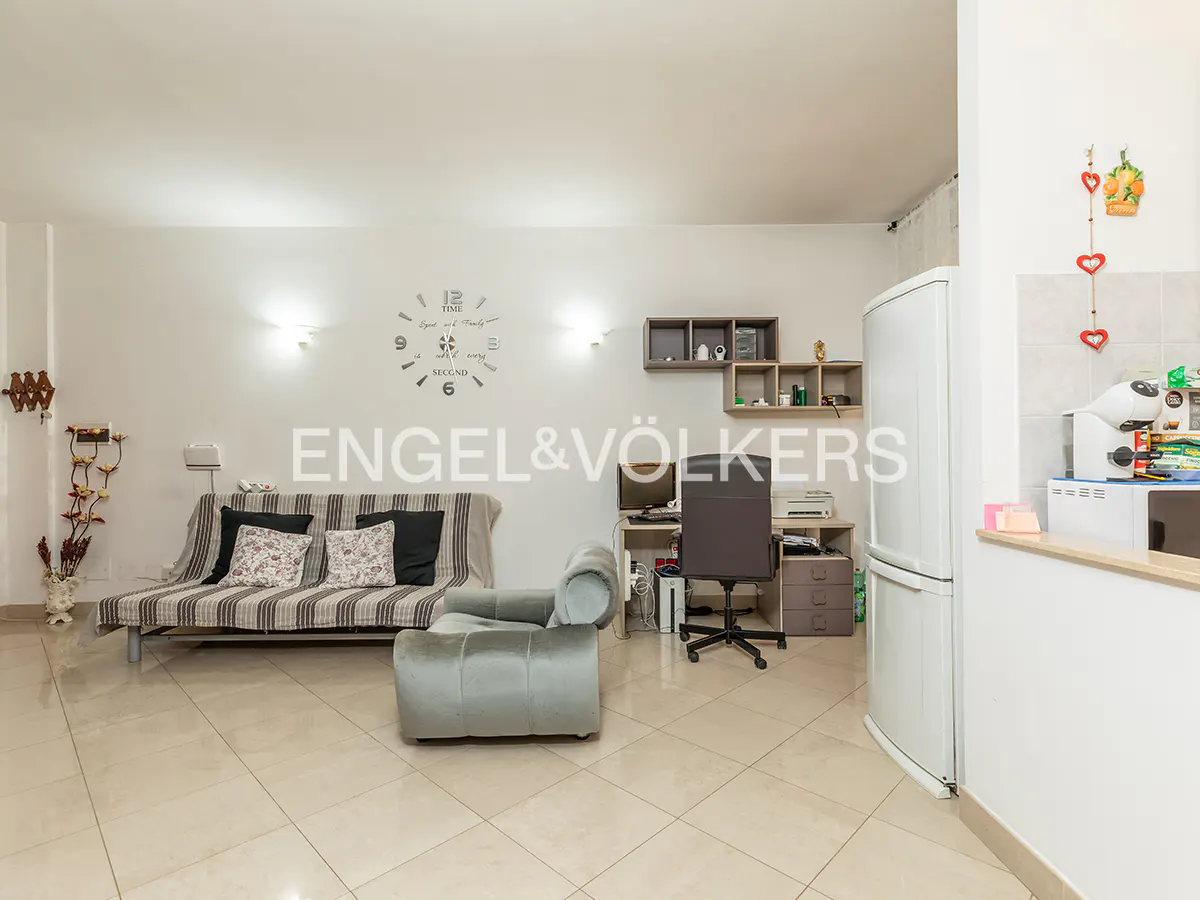 A bright living space with a sofa, chair, desk, and refrigerator. The walls are white, and the floor is tiled.